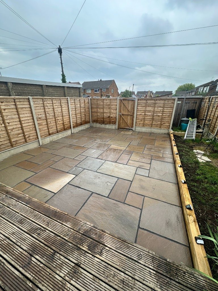 A large riven Indian sandstone patio with dark jointing and timber edging, enclosed by featheredge fencing beside a deck